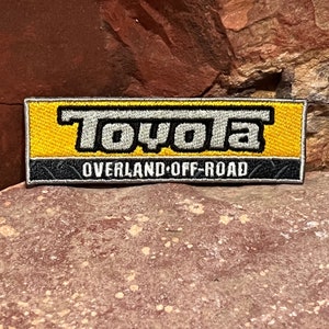 May include: A yellow and gray embroidered patch with the word "Toyota" in gray and the text "Overland Off-Road" in black.