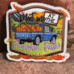 以下が含まれることがあります： 青いピックアップトラックにカボチャが積まれた刺繍パッチ。トラックには「パンプキンパッチで会いましょう」と書かれた看板があります。パッチはオレンジとゴールドの葉で囲まれています。