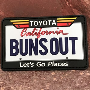 Puede incluir: Un parche de placa de matrícula blanco y negro con el texto "TOYOTA California BUNS OUT Let's Go Places". El parche tiene un diseño rojo, amarillo y naranja en la parte superior.