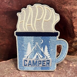 Op de afbeelding: Gestikt patch van een blauwe mok met witte stoom die "Happy Camper" zegt en een kampeerscène in de mok.