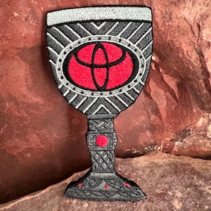 May include: Black and silver embroidered patch of a goblet with a red Toyota logo inside. The goblet has a detailed design with a black and silver pattern.