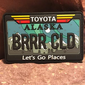 May include: Black and white patch with a blue, green, and white background. The patch features a stylized license plate with the text "TOYOTA ALASKA BRRR CLD" and the words "Let's Go Places".