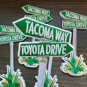 May include: Green and white fabric patches with the text "Tacoma Way" and "Toyota Drive" on them. The patches are attached to a white stick with a green plant at the bottom.