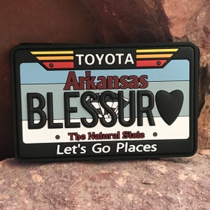 Puede incluir: Parche blanco y negro con un diseño de placa de matrícula. Dice "Arkansas Blessed" con un diamante y un corazón gráfico. El texto "The Natural State" y "Let's Go Places" también está incluido.