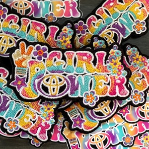 May include: A collection of colorful embroidered patches with the text "Girl Power" in a rainbow font. The patches are decorated with flowers and peace signs.