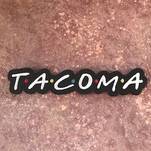 Puede incluir: Parche de fieltro negro con letras bordadas en blanco que deletrean "T.A.C.O.M.A." con puntos de colores entre cada letra.