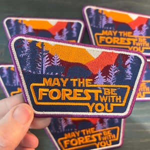 May include: Embroidered patch with a forest scene in shades of blue, orange, and purple. The text "MAY THE FOREST BE WITH YOU" is in a yellow font. The patch has a purple border and is rectangular with rounded corners.