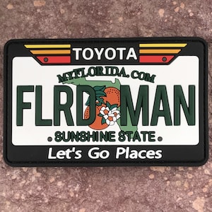 May include: Black and white patch with a stylized Florida license plate design. The plate reads "FLRDMAN" with the text "MYFLORIDA.COM" above and "SUNSHINE STATE" below. The plate is surrounded by a black border with the text "TOYOTA" and "Let's Go Places" in white.