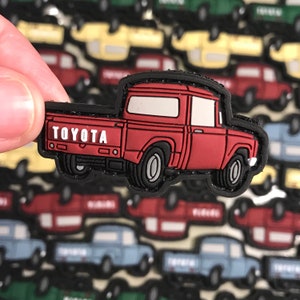 Puede incluir: Un parche de camioneta Toyota roja con un logotipo "TOYOTA" blanco. El parche está hecho de goma y tiene un contorno negro.