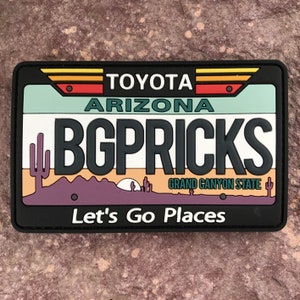 Könnte beinhalten: Ein schwarz-weißes Patch mit einem stilisierten Bild einer Wüstenlandschaft. Das Patch hat den Text "TOYOTA", "ARIZONA", "B.G. PRICKS", "GRAND CANYON STATE" und "Let's Go Places".