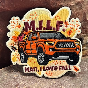 May include: An orange Toyota pickup truck with a bed full of pumpkins. The truck has the words "M.I.L.F." above it and "Man, I Love Fall" below it. The truck is surrounded by fall-themed leaves and flowers.