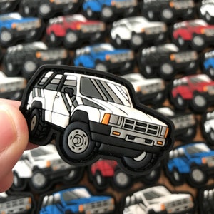 Puede incluir: Un parche blanco, negro y gris de un SUV Toyota 4Runner. El parche está hecho de un material similar al caucho y tiene un diseño detallado.