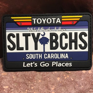 Puede incluir: Un parche blanco y negro con un diseño de placa de matrícula de Carolina del Sur. La placa dice "SLTY BCHS" con un palmetto y una luna creciente. El texto "SOUTH CAROLINA" está debajo de la placa. El texto "TOYOTA" está en la parte superior de la placa con un diseño de rayas rojas, amarillas y naranjas. El texto "While I Breathe, I Hope." está por encima de la placa. El texto "Let's Go Places" está debajo de la placa.