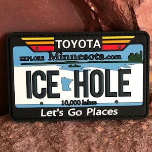 Könnte beinhalten: Ein schwarz-weißes Patch mit einem blauen, weißen und grünen Minnesota-Kennzeichen-Design. Das Kennzeichen lautet "ICE HOLE" mit einem kleinen Umriss des Bundesstaates Minnesota. Oben auf dem Kennzeichen steht "Explore Minnesota.com" und "TOYOTA" mit einem rot-gelb-orangenen Logo. Unten auf dem Patch steht "10.000 Seen" und "Let's Go Places".