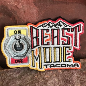 Op de afbeelding: Een geborduurde zwarte en witte patch met de tekst "BEAST MODE TACOMA" in rood, geel en oranje. De patch heeft ook een gestileerde schakelaar met de woorden "ON" en "OFF" in geel.
