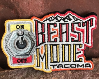May include: A black and white embroidered patch with the text "BEAST MODE TACOMA" in red, yellow, and orange. The patch also features a stylized toggle switch with the words "ON" and "OFF" in yellow.