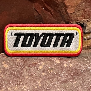 Puede incluir: Parche bordado con la palabra «TOYOTA» en negro, delineada en un rectángulo blanco y bordeada por amarillo y rojo. El parche tiene forma rectangular con esquinas redondeadas.