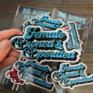 May include: A set of five black and white embroidered patches with the text "Female Owned & Operated" in blue lettering. The patches are on a clear plastic background.