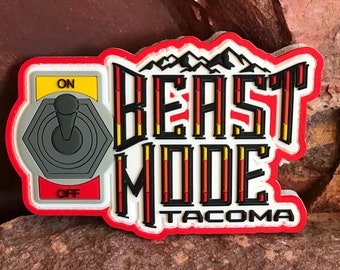 May include: Red, white, and black patch with the words "BEAST MODE" and "TACOMA" in a stylized font. The patch features a toggle switch with the words "ON" and "OFF" and a mountain range graphic.