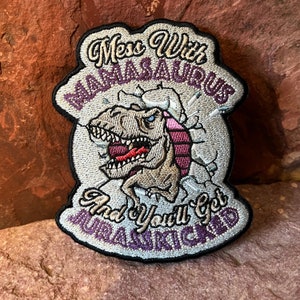 Puede incluir: Un parche bordado blanco y morado con un dinosaurio de dibujos animados rompiendo una pared. El texto en el parche dice "Mess with a Mamasaurus and you'll get Jurassic'd".