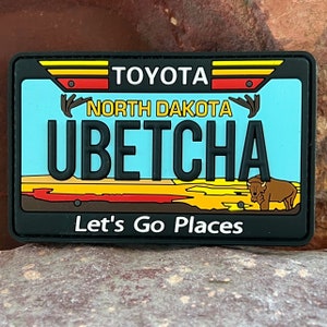 Könnte beinhalten: Ein schwarzer und gelber Nummernschild-Patch mit dem Text "TOYOTA", "NORTH DAKOTA" und "UBETCHA". Der Patch zeigt außerdem eine rote, gelbe und braune Landschaftsszene mit einem Bison in der unteren rechten Ecke. Der Text "Let's Go Places" ist unter dem Nummernschild gedruckt.