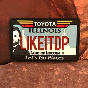 Può includere: Toppa bianca e nera con un design di targa blu, bianca e rossa. La targa recita "TOYOTA ILLINOIS LIKE IT DP LAND OF LINCOLN Let's Go Places".