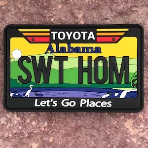 Può includere: Un cerotto per targa dell'Alabama nero e giallo con il testo "SWT HOM" e le parole "Toyota" e "Let's Go Places".