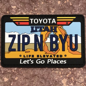 Könnte beinhalten: Ein schwarzer und gelber Patch mit einem Utah-Kennzeichen-Design. Das Kennzeichen lautet "ZIP N BYU" mit den Worten "TOYOTA" und "LIFE ELEVATED" darüber und darunter. Der Text "Let's Go Places" befindet sich unter dem Kennzeichen.