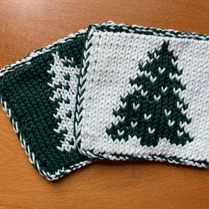 May include: Two square knitted coasters featuring a white pine tree design on a dark green background and a dark green pine tree design on a white background. Each coaster has a braided white and green border edge with a textured knit stitch.