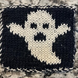 Puede incluir: Un cuadrado tejido con un diseño de fantasma blanco sobre un fondo negro. El fantasma tiene una cara simple y sonriente. Los bordes están acabados con un borde blanco, creando un artículo decorativo con temática de Halloween.