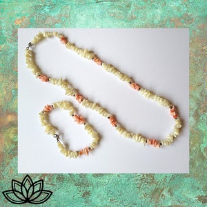 May include: A necklace and bracelet set made from cream-coloured shell chips and coral-coloured beads. The jewellery has small, dark metal accents. The set is displayed on a white surface with a textured green background.