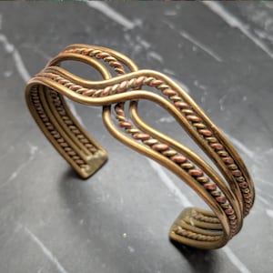 May include: A copper-toned cuff bracelet with a woven design. The bracelet features a mix of copper and bronze tones, with a textured, braided pattern. The open cuff design allows for easy wear. The bracelet is displayed on a dark, marbled surface.