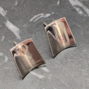 May include: A pair of silver-toned stud earrings featuring a smooth, curved, rectangular metal surface. Each earring has a polished, reflective finish that catches the light, creating a subtle gradient effect across the slightly concave metal shape.