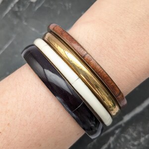 May include: A set of four stacked bangle bracelets worn on the wrist. The stack includes a dark brown polished bangle, a smooth off-white bangle, a metallic gold-toned bangle with a reflective finish, and a reddish-brown wood-grain textured bangle.