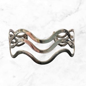 May include: A silver-toned cuff bracelet featuring three parallel, interconnected wavy bands. The metal has a polished, reflective finish, and the design consists of continuous, flowing curves that create an open, airy structure throughout the piece.