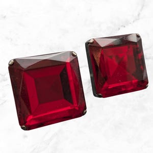 May include: Two square-cut, deep red gemstone earrings. Each earring has a faceted, rectangular gem set in a silver-toned metal frame. The gems have a rich, transparent appearance, reflecting light. The earrings are displayed on a white marble surface.