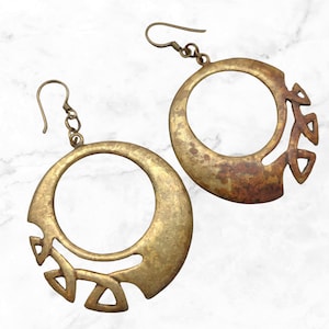 May include: A pair of brass-coloured hoop earrings. Each earring has a circular design with cut-out geometric shapes. The earrings have a hook closure and show signs of wear, giving them a vintage appearance.
