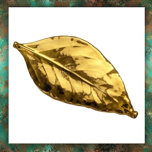 May include: A gold-coloured leaf ornament with intricate veining and a shiny surface. The leaf is set against a white backdrop, framed by a textured green and brown border. The design is elegant and inspired by nature. The object is likely for decorative purposes.
