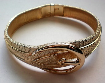 May include: A gold-tone metal bracelet with a buckle design. The bracelet is textured and has a shiny finish.