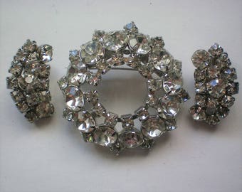 Può includere: Un set di tre gioielli in strass color argento. Il pezzo centrale è una grande spilla circolare con un anello di strass. I due pezzi più piccoli sono orecchini con un gruppo di strass.