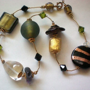 Puede incluir: Un collar de cuentas de vidrio dorado, negro y verde con una variedad de formas y tamaños. El collar presenta cuentas cuadradas, redondas y rectangulares, así como algunas cuentas más pequeñas entre ellas.