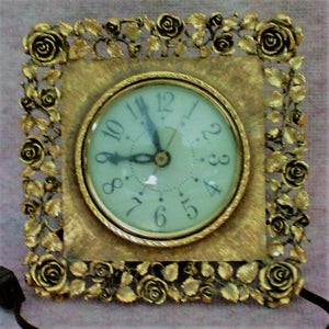 May include: Gold-toned clock with a white face and black hands. The clock is surrounded by a decorative frame with roses and leaves.