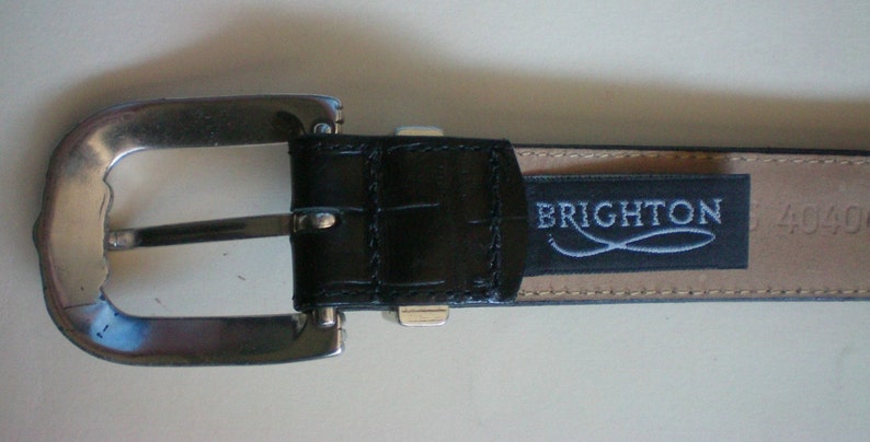 Peut inclure: Une ceinture en cuir noir avec une boucle argent&eacute;e et un logo Brighton. La ceinture a un motif crocodile et le logo est emboss&eacute; en argent.