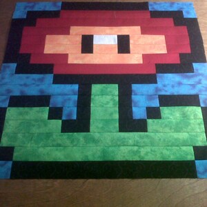 May include: A red and green pixelated flower with a black outline. The flower is made of fabric and has a blue background.