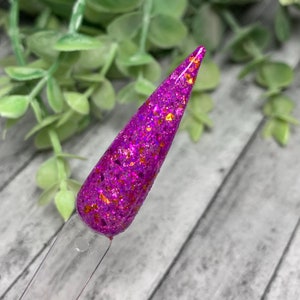 May include: A close-up of a purple nail polish with gold glitter. The polish is applied to a nail tip.