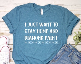 以下が含まれることがあります： 白地に「I just want to stay home and diamond paint」と書いてあるターコイズブルーのTシャツ。Tシャツは白い星の列で飾られています。
