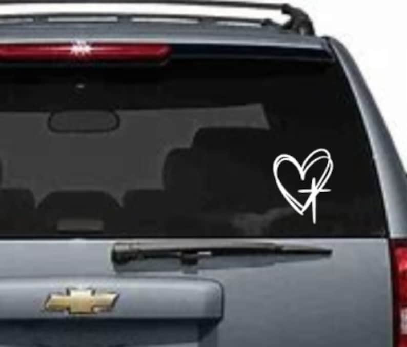 May include: White vinyl decal of a heart with a cross inside. The decal is on the back window of a gray SUV.