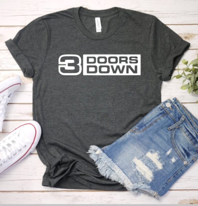May include: Dark gray t-shirt with the text "3 DOORS DOWN" in white block letters. The shirt is paired with denim shorts and white sneakers. The t-shirt is made of a soft material.