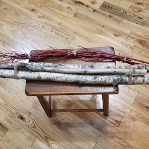 May include: Birch logs with natural bark and twine, alongside a bundle of red twigs, rest on a small wooden stool. The logs have a white and black pattern. The red twigs add a pop of color to the natural wood tones.