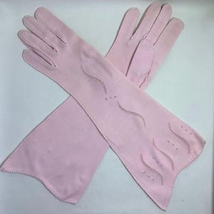 May include: A pair of pale pink gloves, likely for formal wear. The gloves are long, extending past the wrist, and feature decorative stitching and small embellishments. The gloves are made of a soft material.
